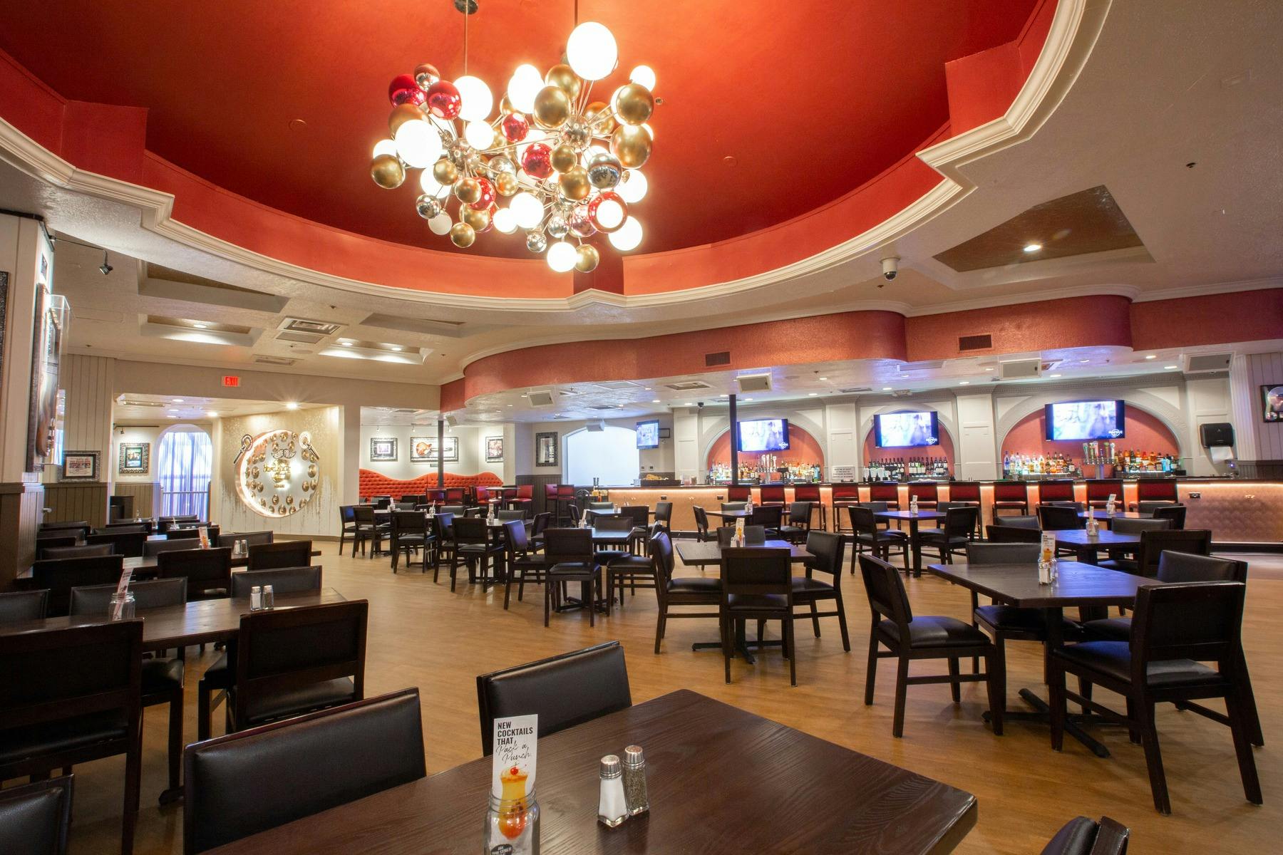 A spacious restaurant with a modern chandelier, red ceiling, black chairs, wooden tables, and a bar with multiple TV screens.