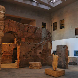 Forntida tegel- och stenruiner inuti en modern byggnad med en partiell staty på en träbas i förgrunden.