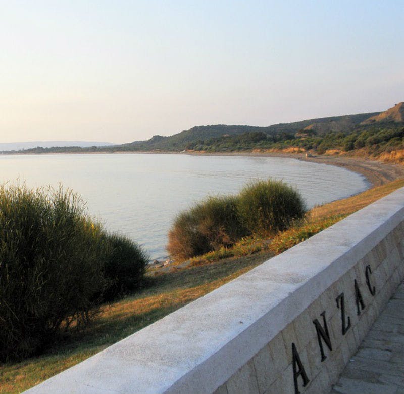 Una playa tranquila con suaves olas, rodeada de vegetación y un muro de piedra con la inscripción "ANZAC". Colinas visibles al fondo.