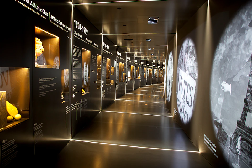 A dimly lit hallway in a museum exhibit displays historical information from the 1900-1910 era, with illuminated text and artifacts.