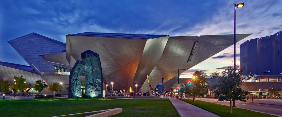 Modern angular building with unique architecture, illuminated at dusk. People are gathered near its entrance and sculptures.