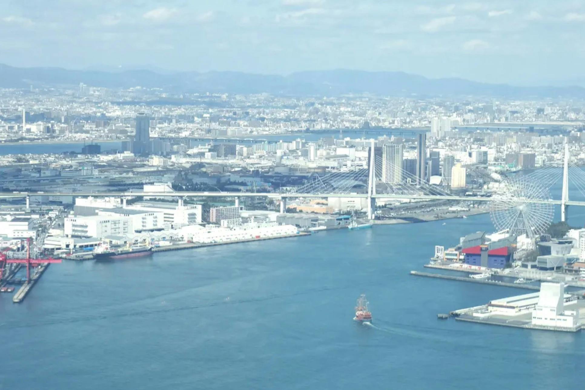 Uma paisagem urbana com uma grande zona ribeirinha, uma ponte suspensa, uma roda gigante, edifícios industriais e um navio na água.