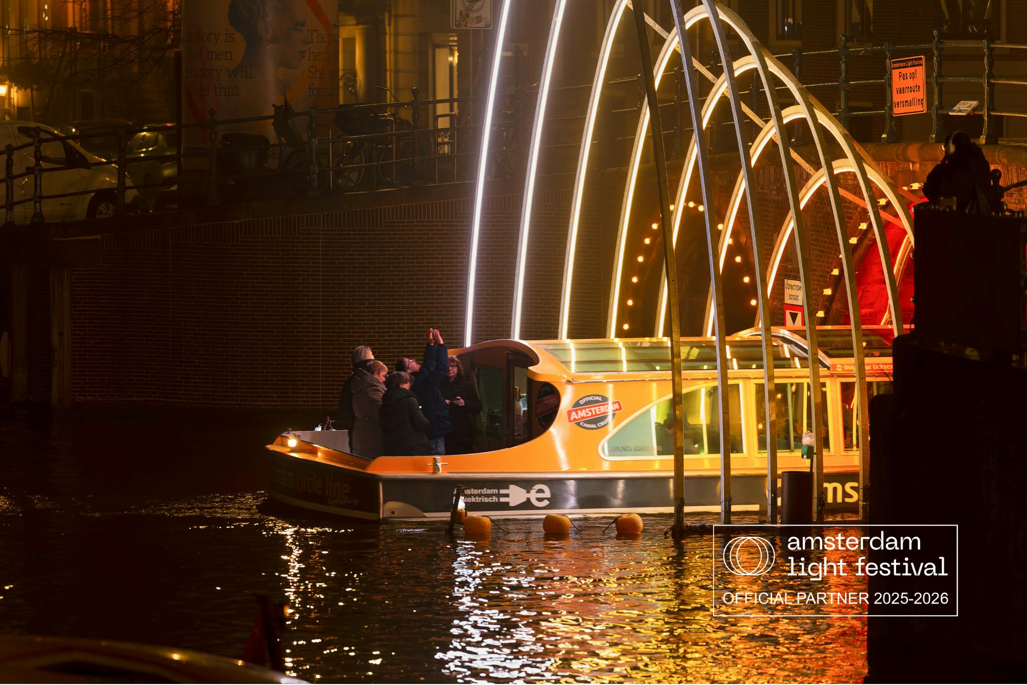 A group of people on a boat pass under illuminated arches at night with "Amstel Light OFFICIAL PARTNER" text in the corner.
