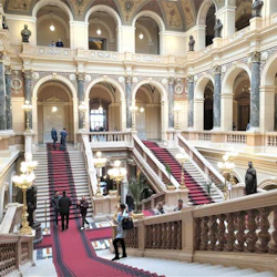 Stor trappa med röda mattan och utsmyckade dekorationer i en historisk byggnad. Människor går och observerar arkitekturen.