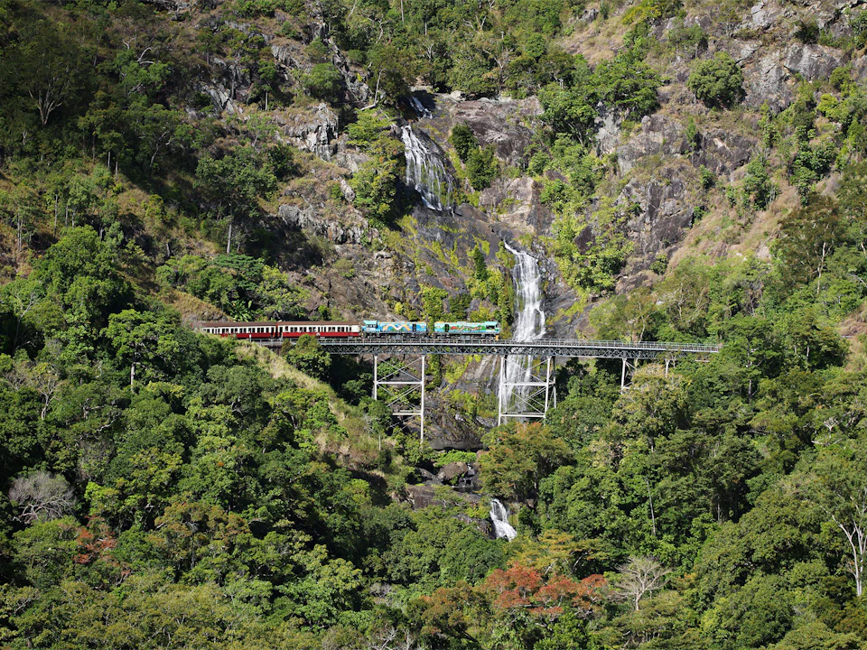 A train travels on a bridge surrounded by lush green forest and a cascading waterfall in the background.