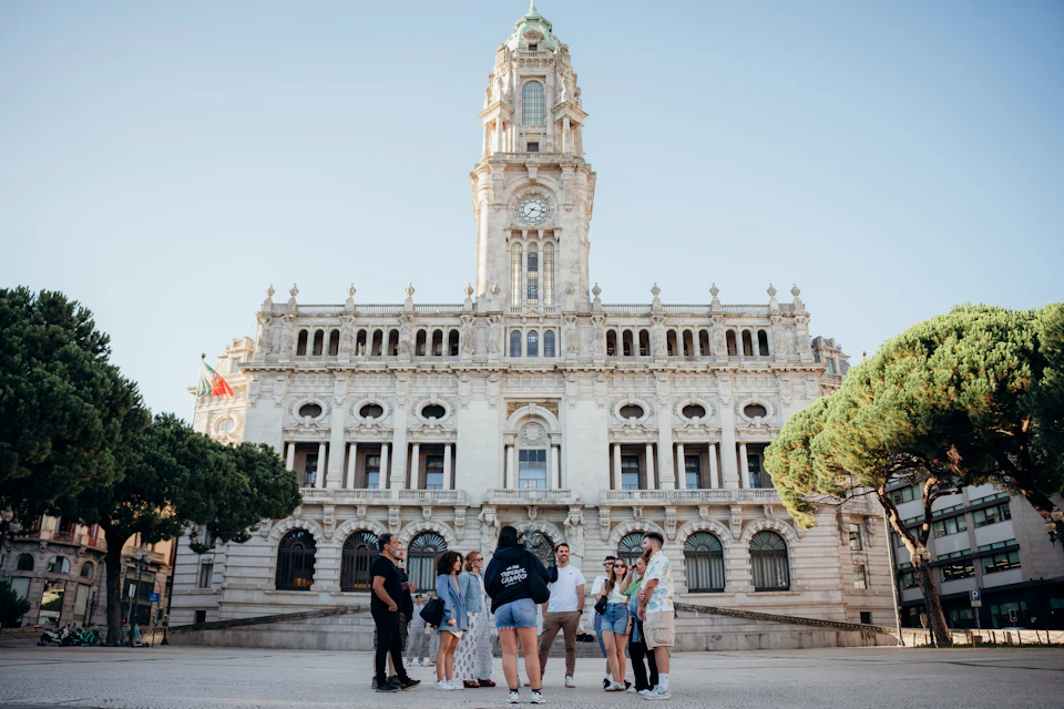 A group of people standing in front of a large historic building with a clock tower, trees, and a clear sky.