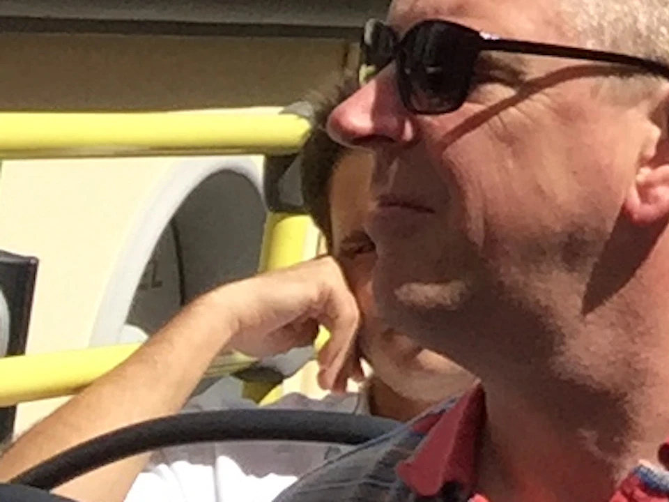 Tourists on an open-top bus tour ride through a narrow street lined with yellow buildings under a blue sky with few clouds.