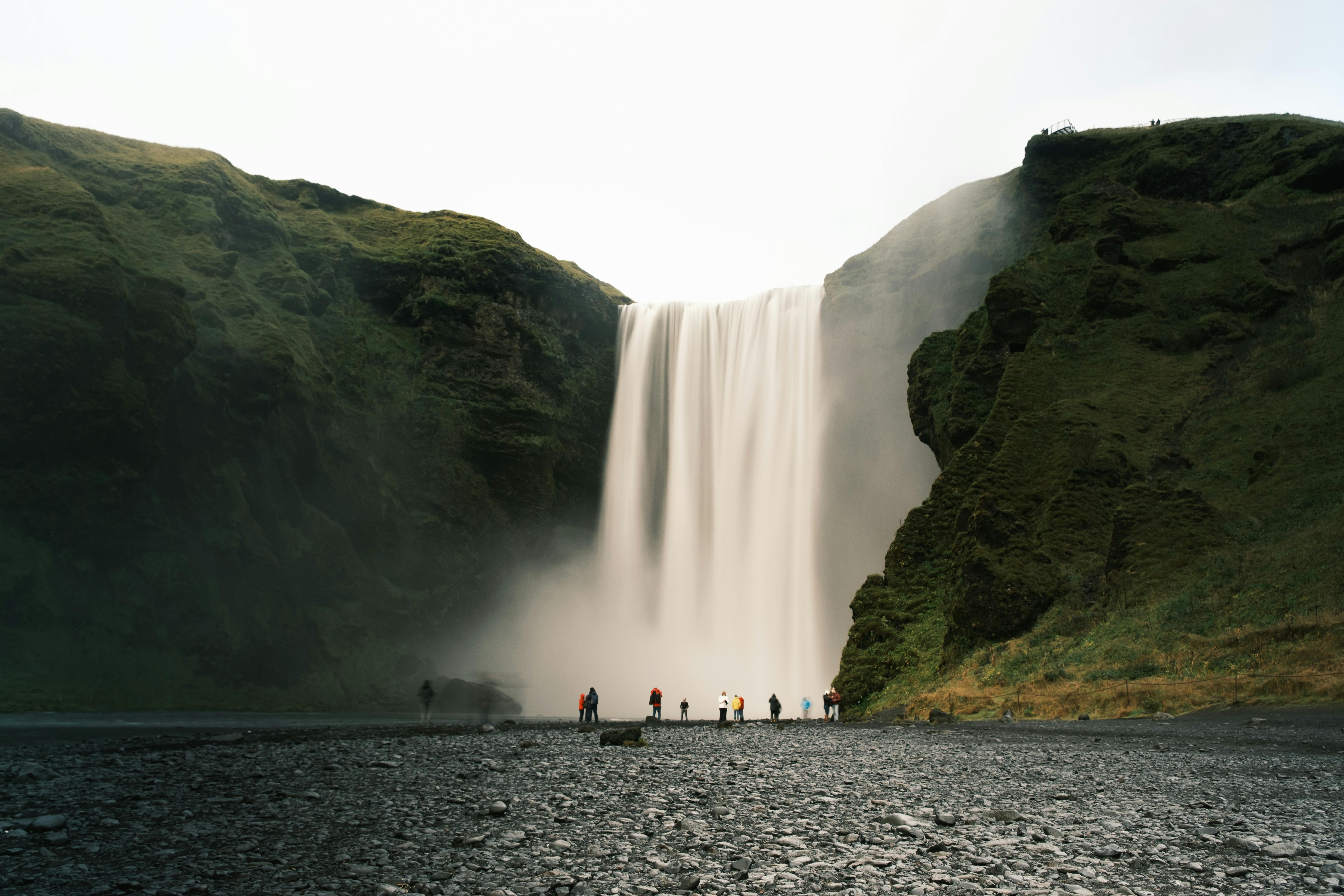 Large waterfall cascading between green cliffs with people standing at the base on a rocky ground, observing from a distance.