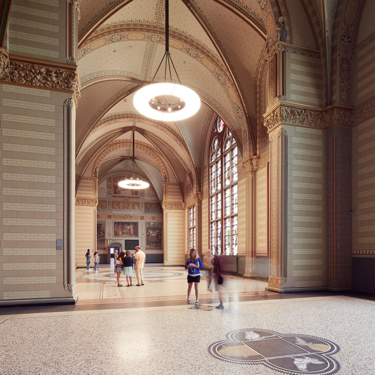 A spacious hall with high vaulted ceilings, decorative details, a large circular light, and people walking or standing.