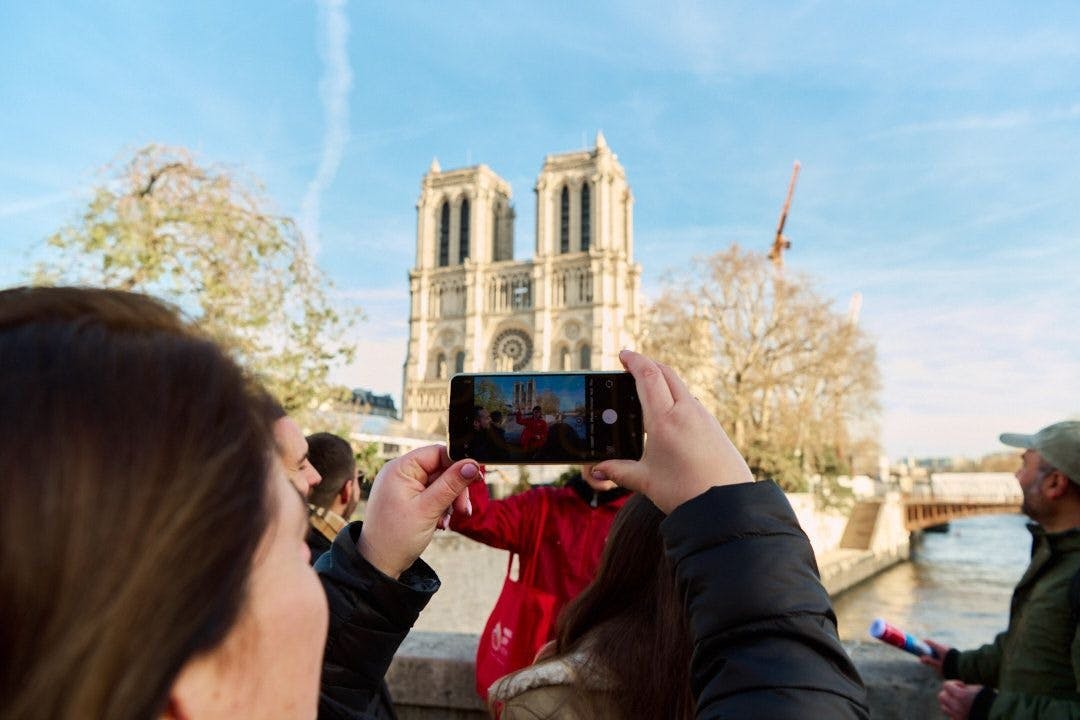 A person holds up a smartphone to take a photo of Notre-Dame Cathedral, with other people and crane visible in the background.