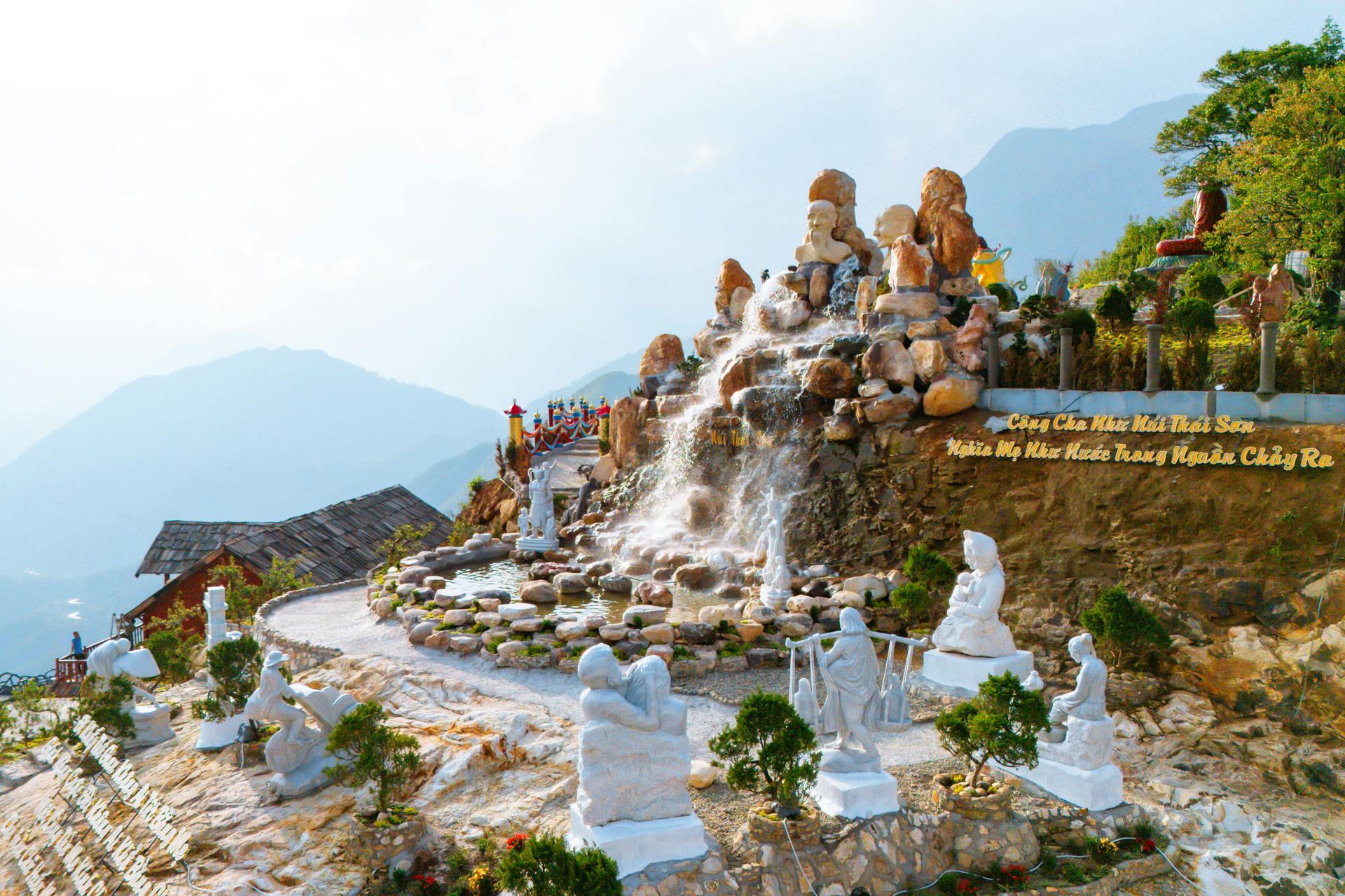 A scenic outdoor area featuring a waterfall cascading over rock formations surrounded by statues and greenery, with distant mountains.