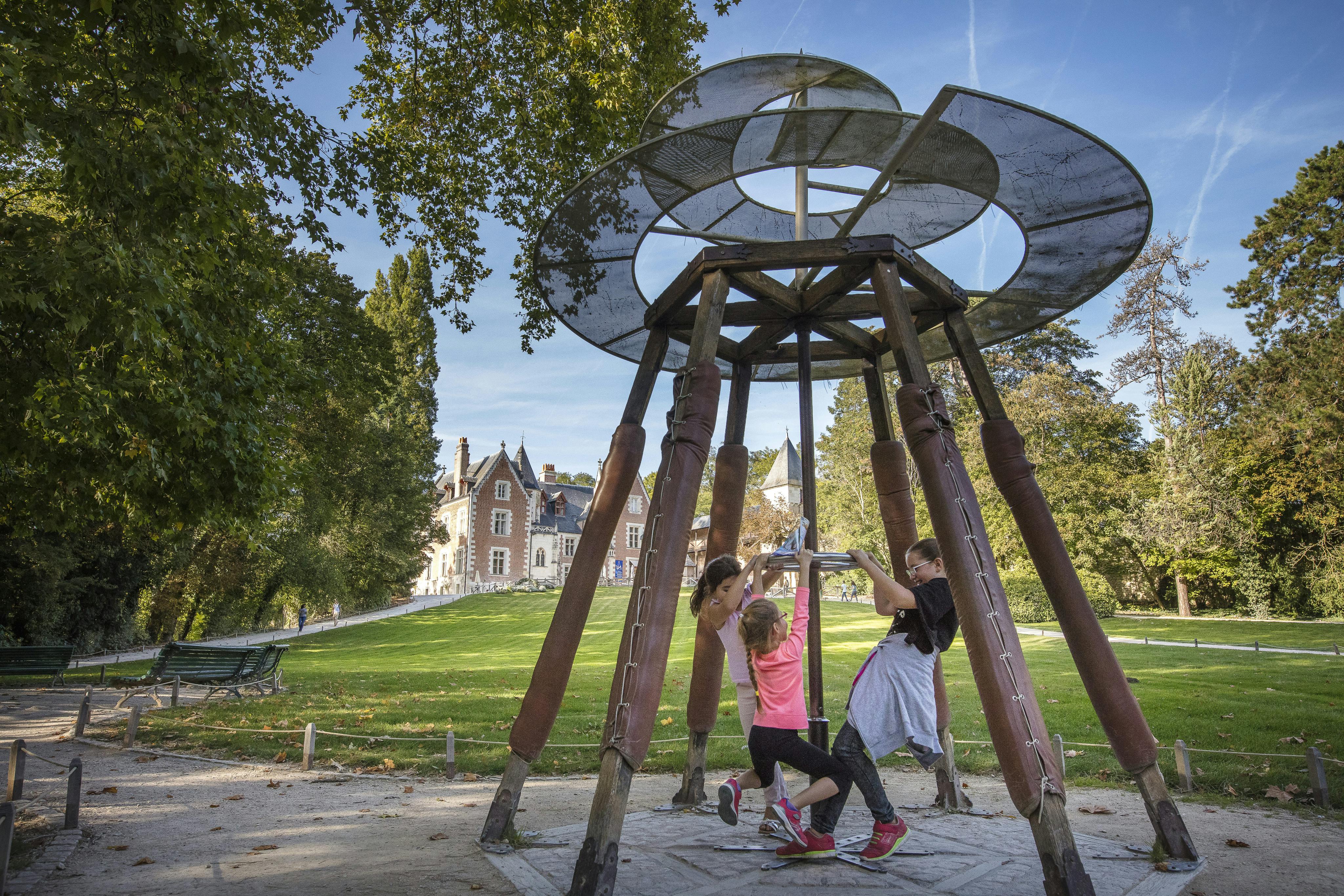 Tres niños juegan en una estructura giratoria de un parque infantil, con un edificio grande y unos árboles al fondo.