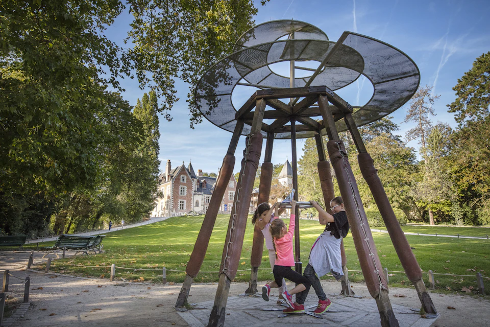 Three children play on a spinning playground structure in a park, with a large building and trees visible in the background.