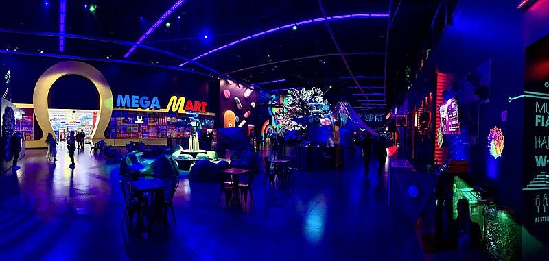 Un'area di divertimento al coperto molto illuminata con un ingresso "Mega Mart", luci al neon, decorazioni futuristiche e persone che camminano e si siedono.