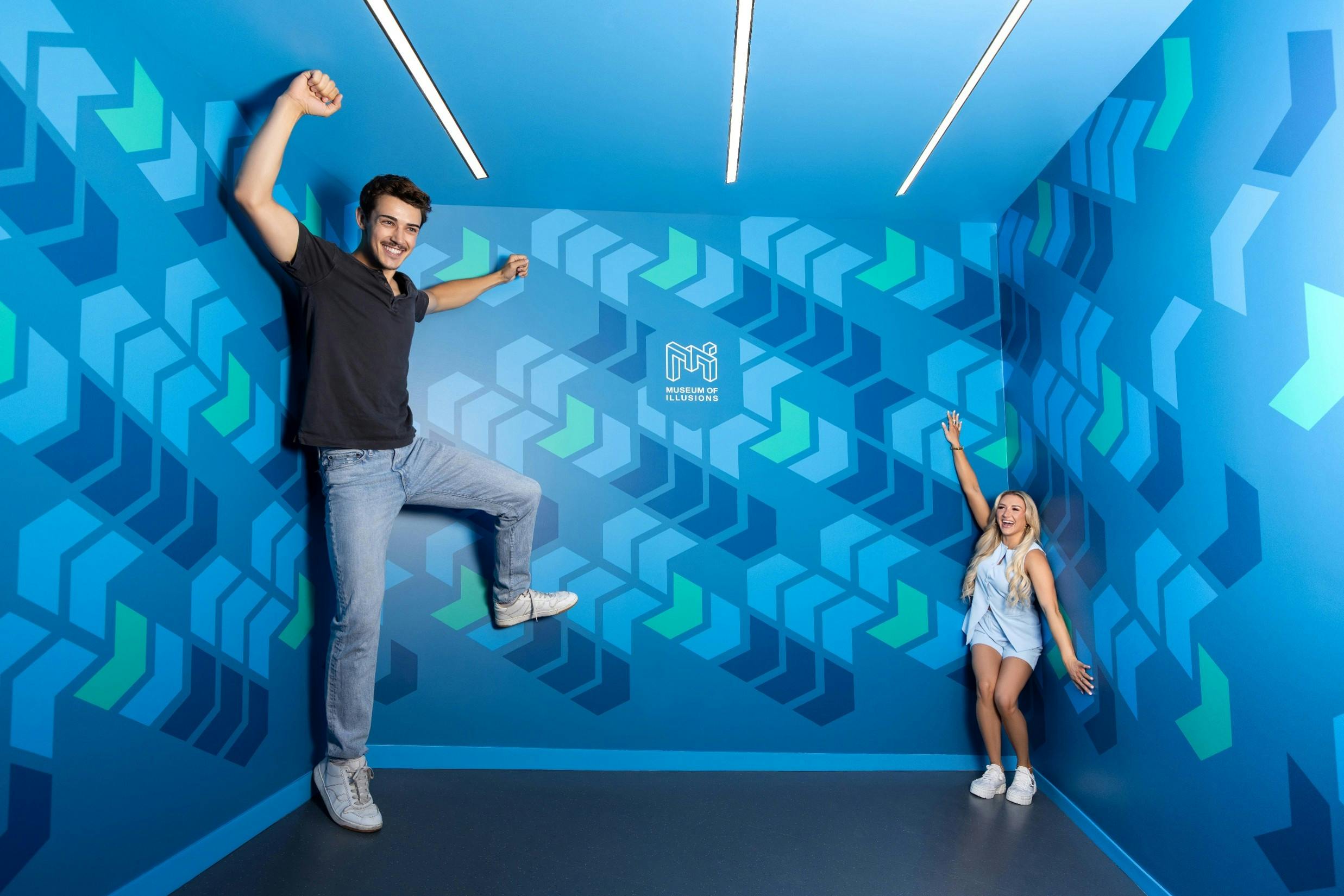 Deux personnes posant dans une pièce bleue avec des motifs géométriques et le logo "Musée des illusions" sur le mur du fond.