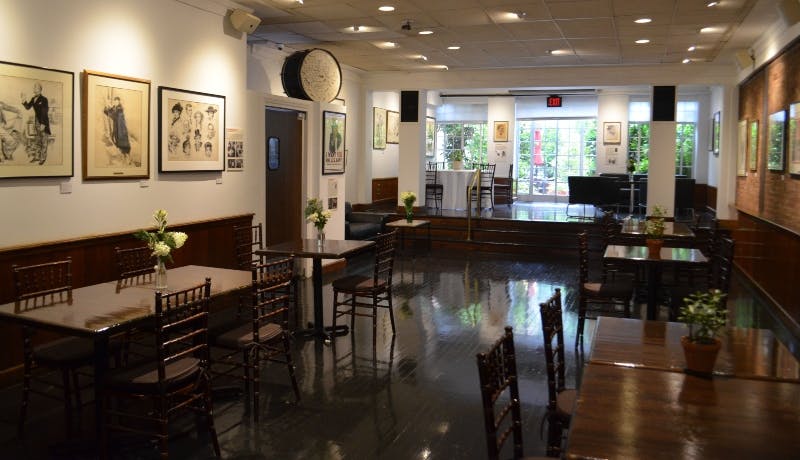A brightly lit cafe with wooden chairs and tables, framed pictures on the walls, and plants near the entrance.