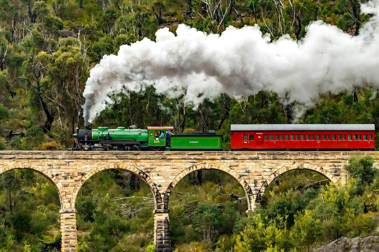 A green steam train with red passenger cars crossing a stone bridge in a forested area, emitting white smoke.