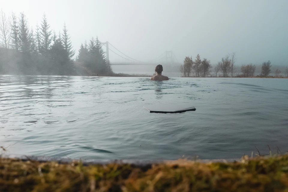 A person in an outdoor pool facing a misty landscape with trees and a bridge in the background.