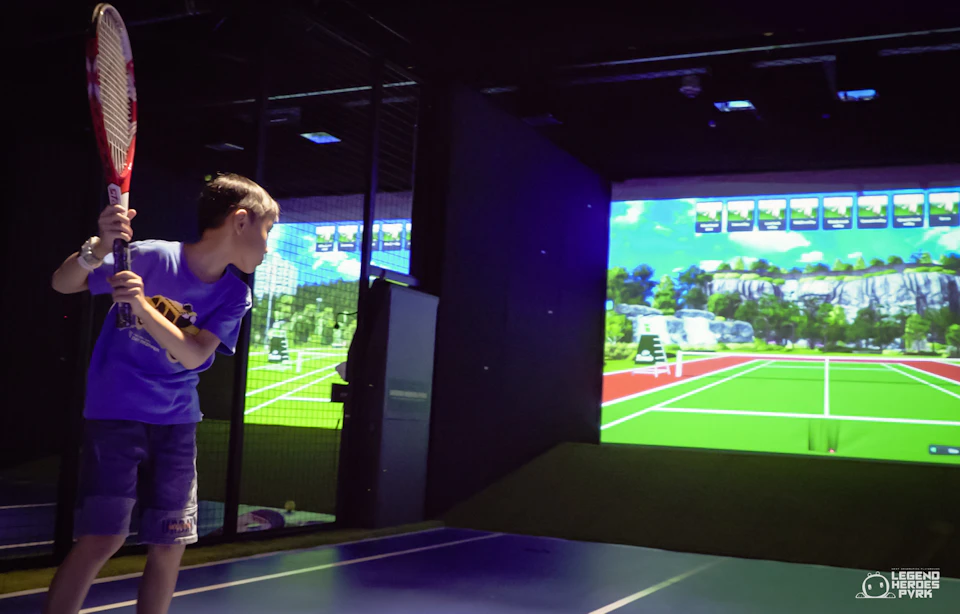 A child holding a toy stands in front of a large screen displaying a virtual tennis court in an indoor setting.