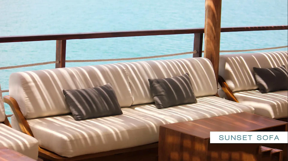 A cushioned outdoor sofa with striped shadows, gray pillows, and wooden railing overlooks calm blue water.