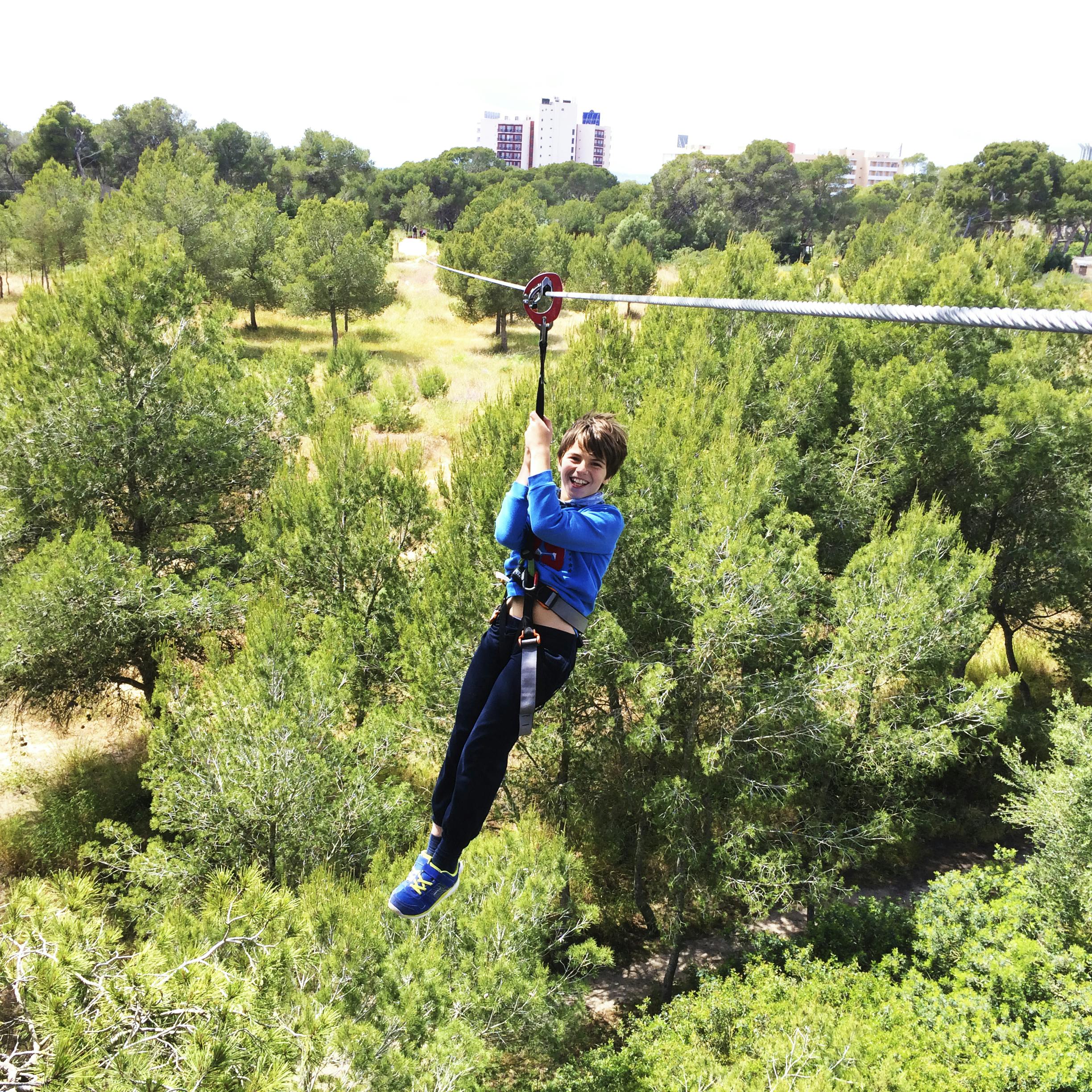 A person zip-lining over a forested area with city buildings in the distant background.