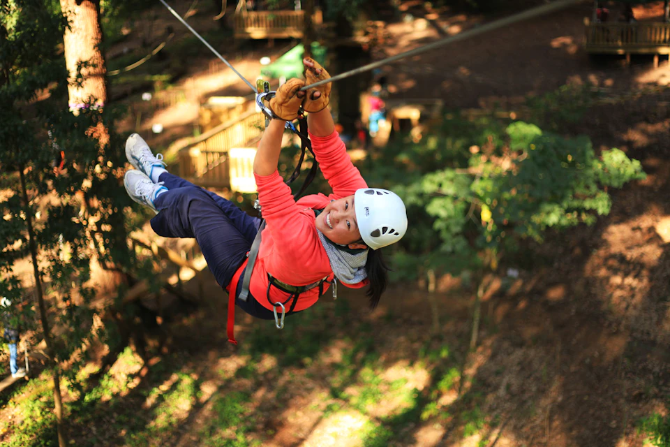 Person wearing a helmet and gloves zip-lining over a forested area, smiling with trees and a wooden platform below.
