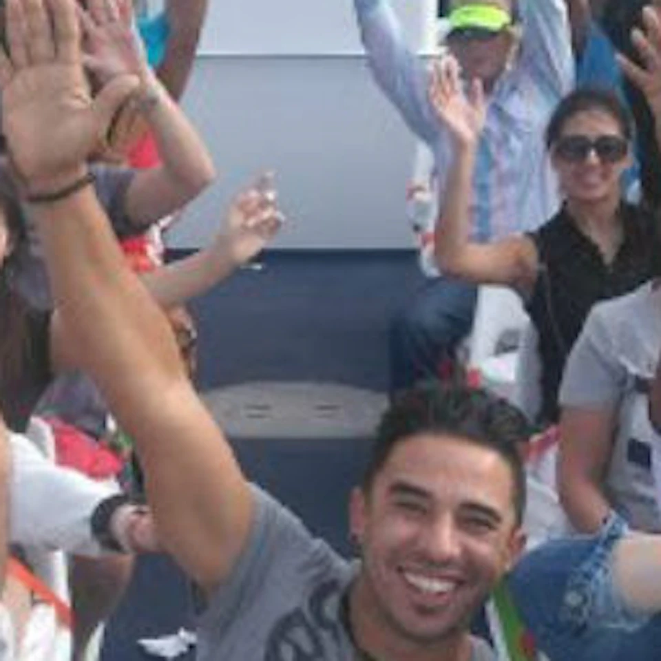 Group of people on a boat ride, smiling and raising their hands, with water and cloudy sky in the background.