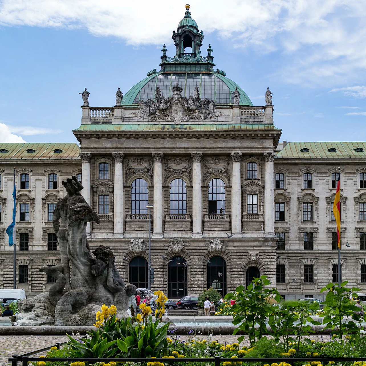 Ornate historic building with arched windows and a green dome, a stone fountain with human figures, and yellow flowers in the foreground.