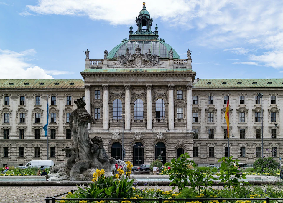 Ornate historic building with arched windows and a green dome, a stone fountain with human figures, and yellow flowers in the foreground.