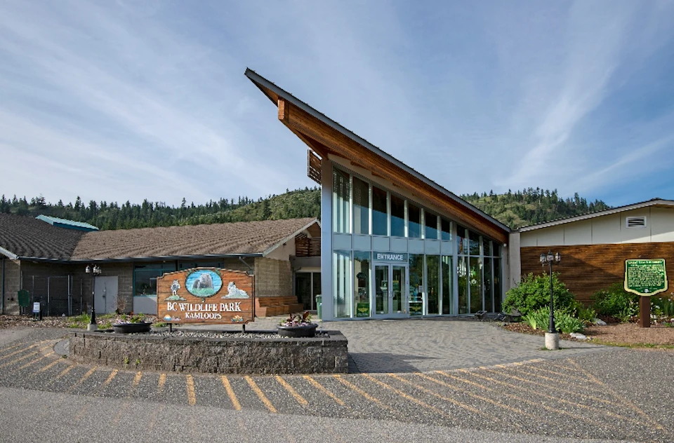 Building entrance with a triangular roof, large glass windows, and a sign reading “BC Wildlife Park Kamloops.” Hill and sky in background.