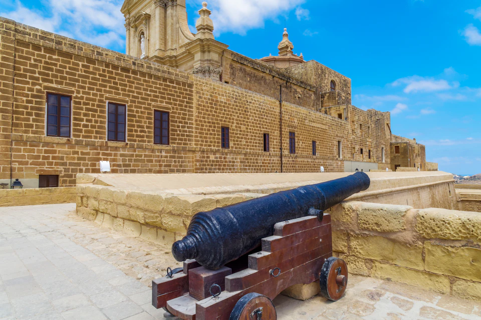A historic stone building under a blue sky, featuring a large black cannon mounted on a wooden carriage in the foreground.