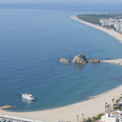 Aerial view of a curved coastline with a ship near the shore, large rocks in the water, sandy beach, and buildings in the distance.
