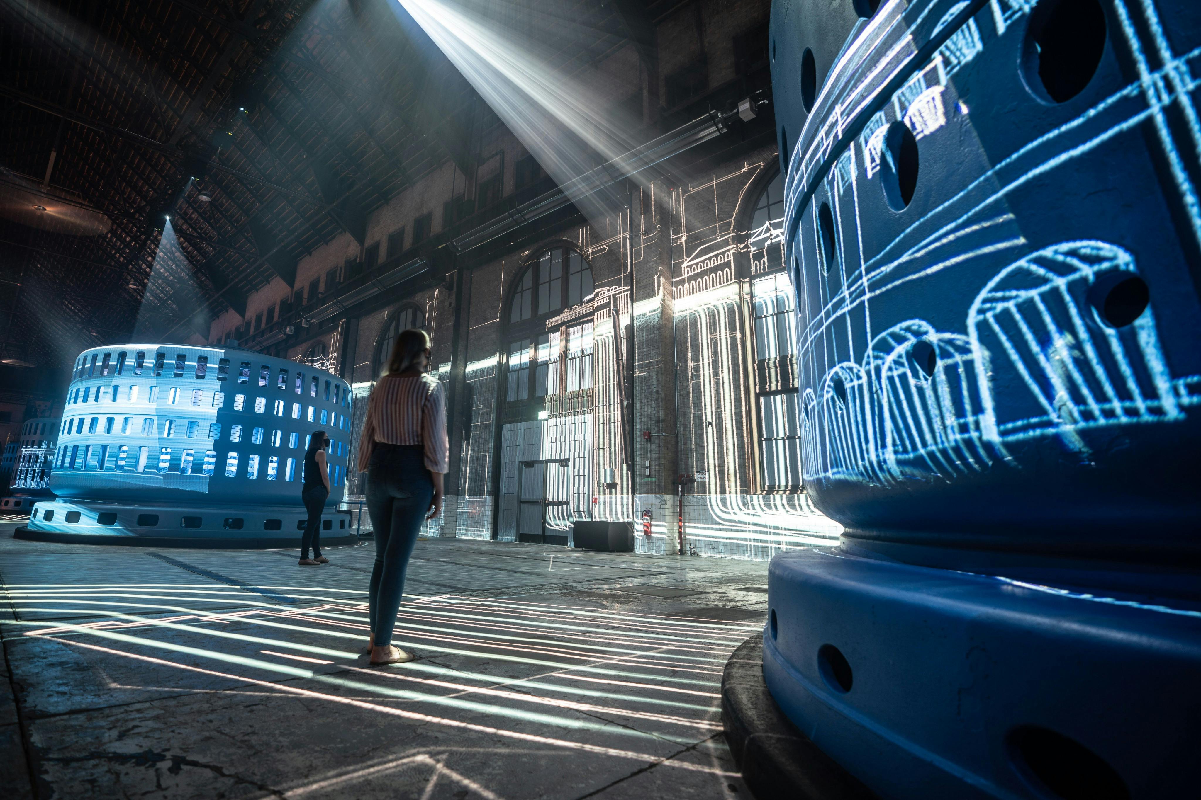 Člověk stojí ve velkém prostoru s architektonickými projekcemi na stěnách a velkými modrými konstrukcemi.