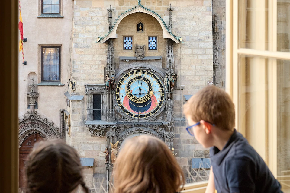 Three children look out a window at an ornate astronomical clock on a historic building facade.