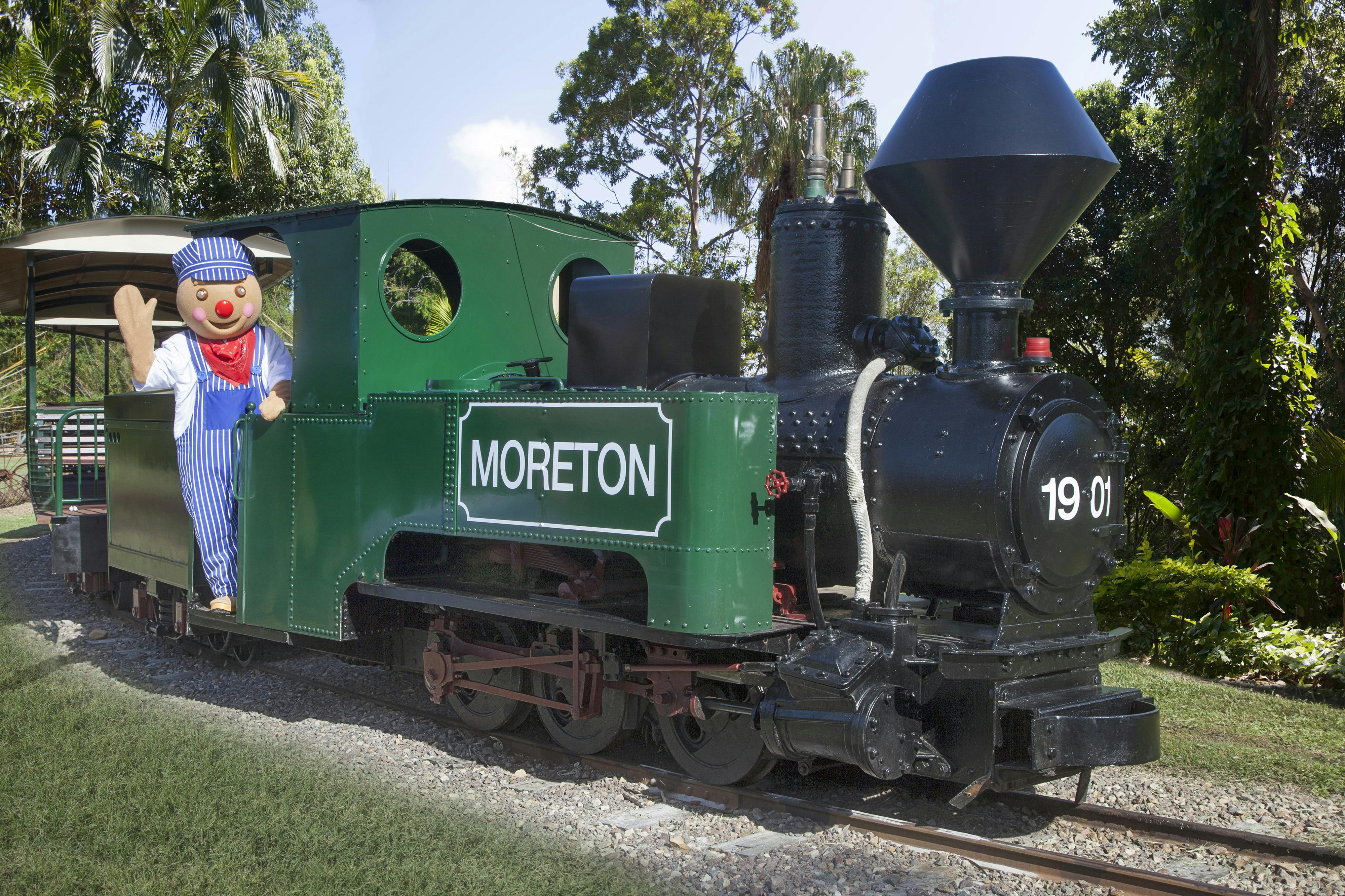 一个穿着火车工程师服装的人站在标有“摩顿”的绿色火车发动机上，周围环绕着树木。