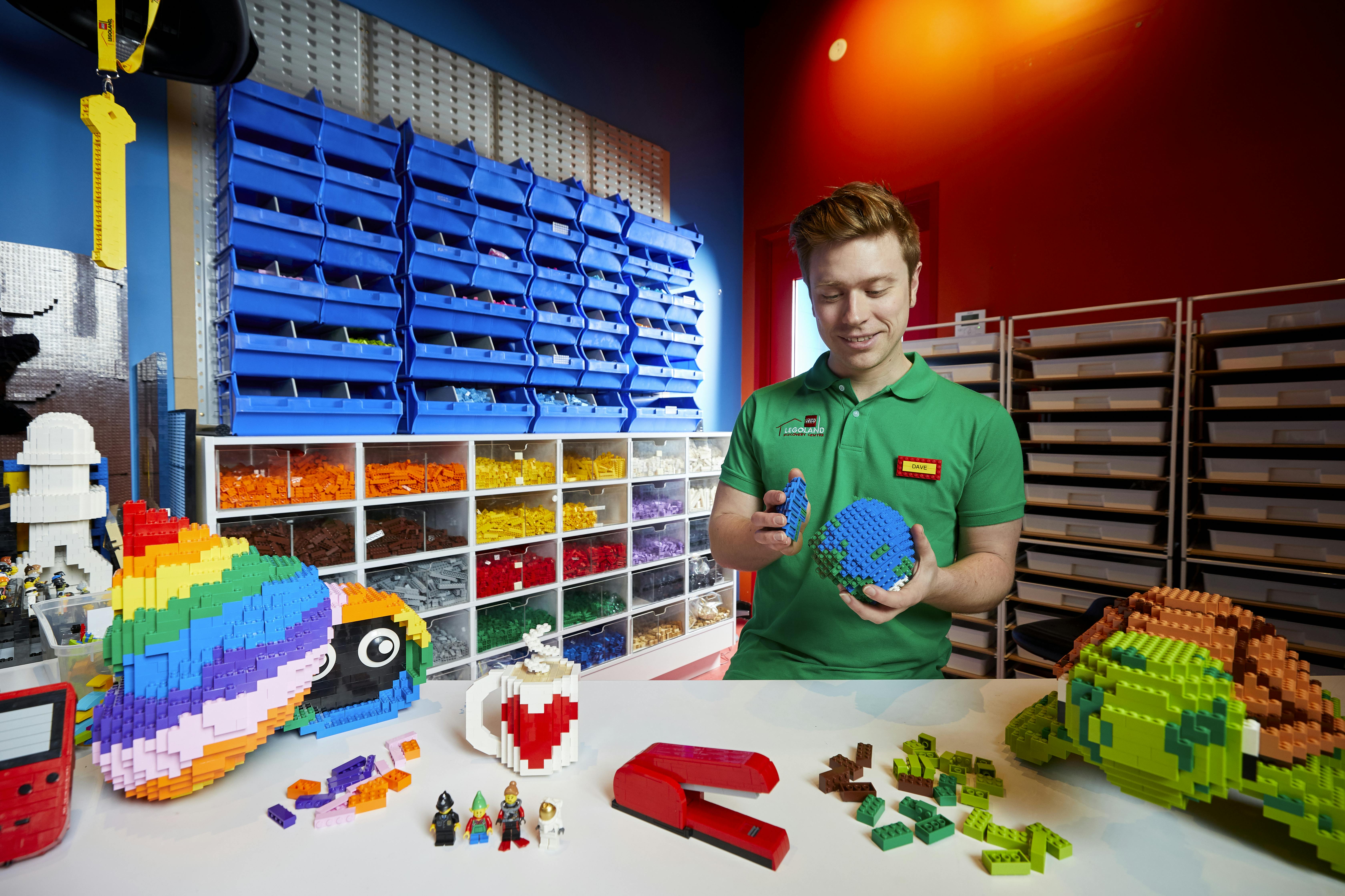 A person in a green shirt holds a LEGO Earth near a table with LEGO models, surrounded by organized LEGO storage bins.