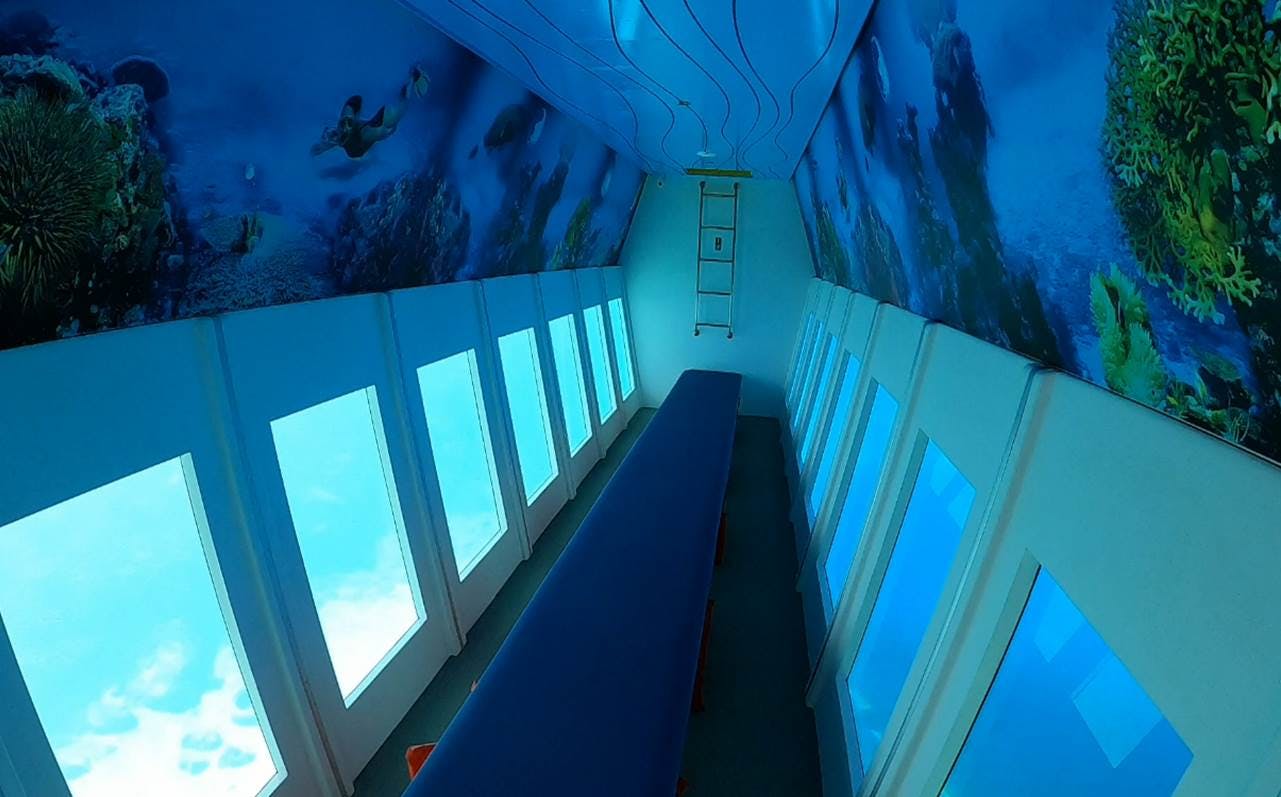 A narrow room with large windows on both sides, blue seating in the center, and underwater-themed decorations on the ceiling and walls.