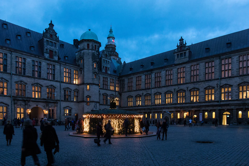 People walking in a cobblestone square with a building adorned with lights in the background and a circular, lighted kiosk.