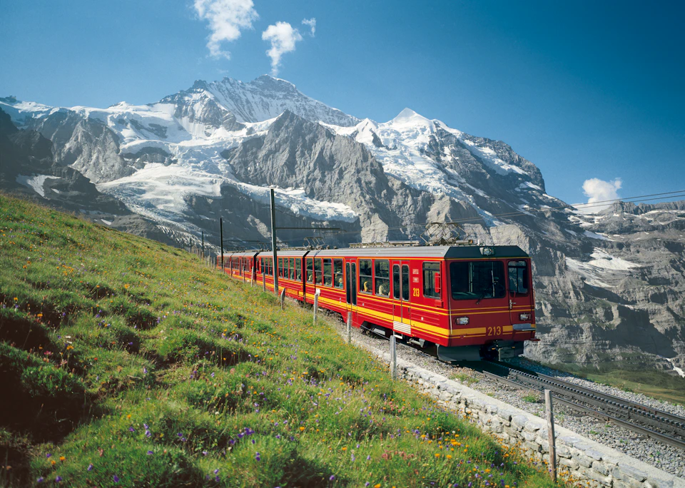 A red and yellow train travels on a mountainside with snow-capped peaks in the background under a clear blue sky.