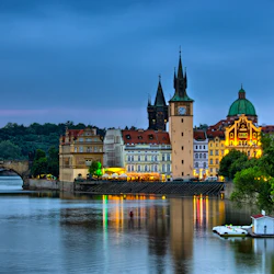 Historisk stadsbild med upplysta byggnader, ett klocktorn och en stenbro som reflekterar över en lugn flod i skymningen.