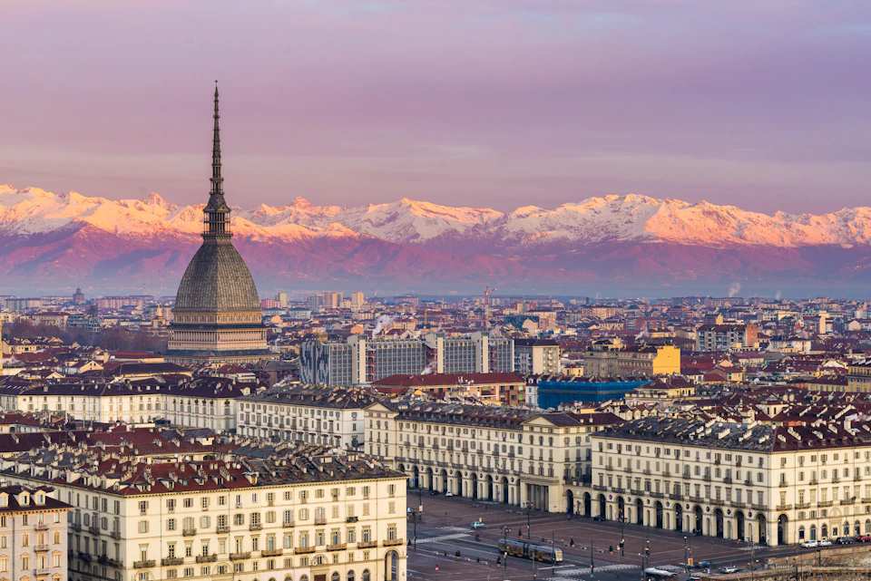 Cityscape with a prominent dome structure, surrounding buildings, and snow-capped mountains under a purple-pink sky.