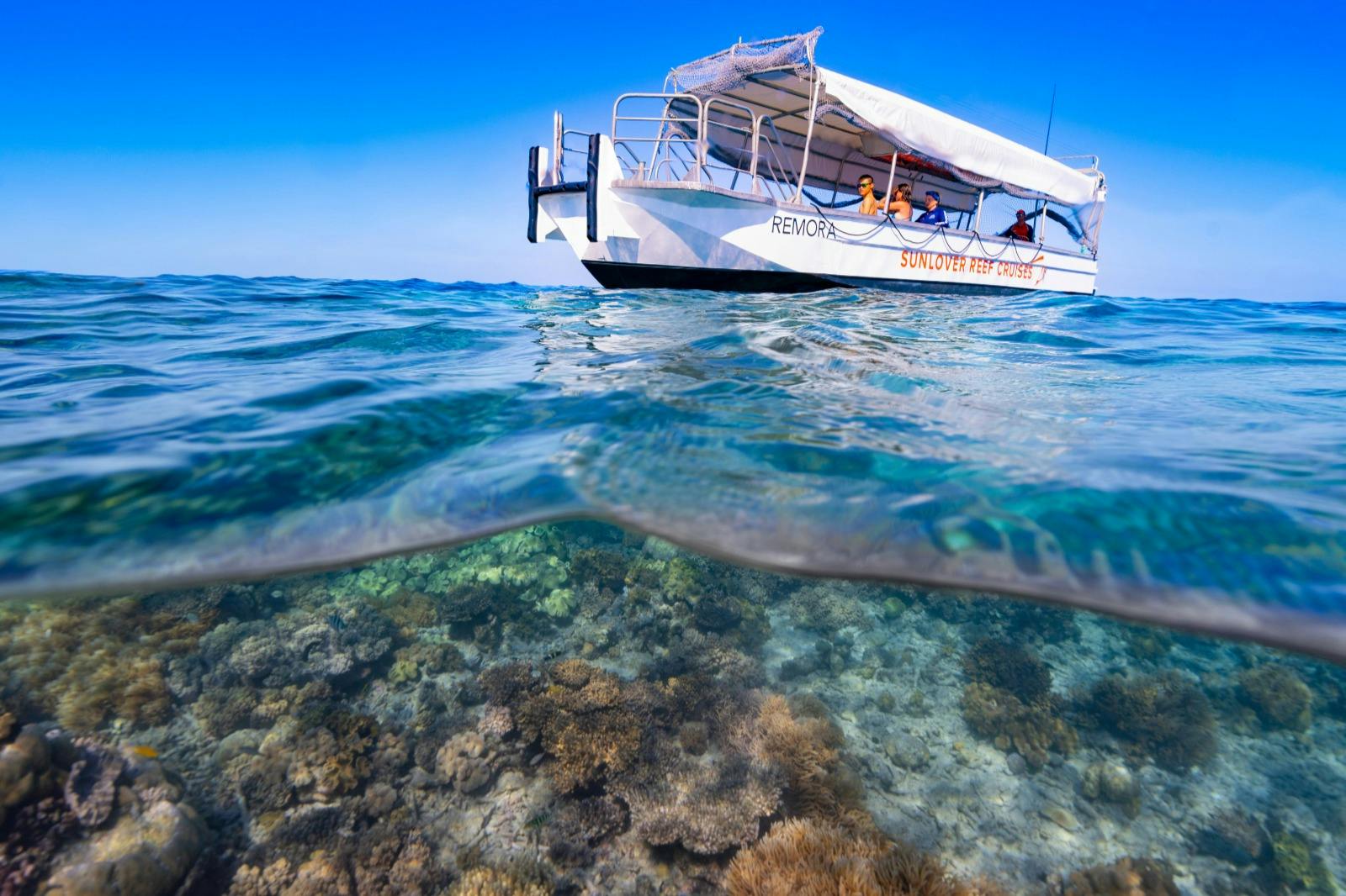 A partially submerged view of a boat with people aboard, floating above a vibrant coral reef in clear blue water.