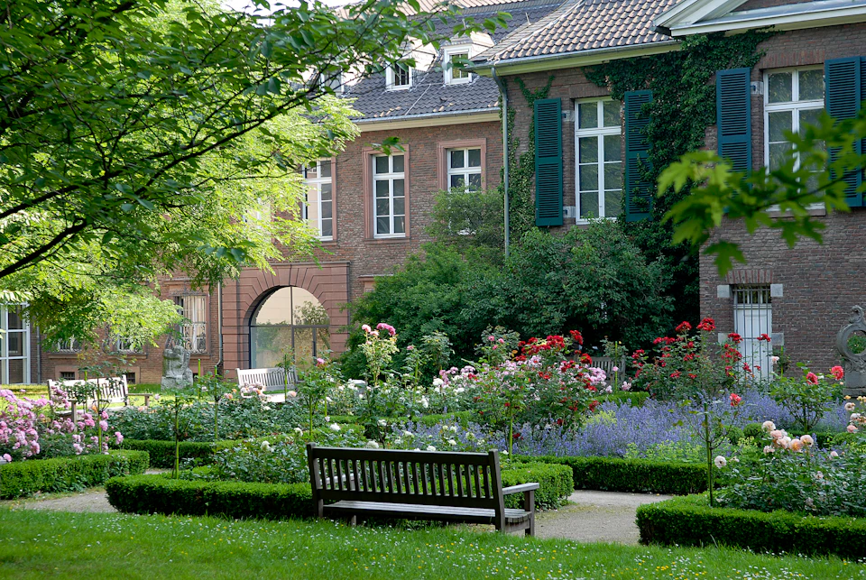 A peaceful garden with flower beds, a wooden bench, and a brick building with shuttered windows partially covered in ivy.