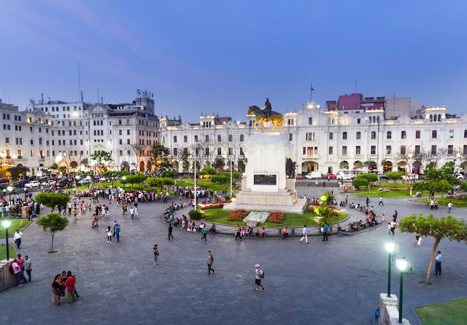 People walking and sitting around a large plaza with a central equestrian statue, surrounded by historic buildings at dusk.