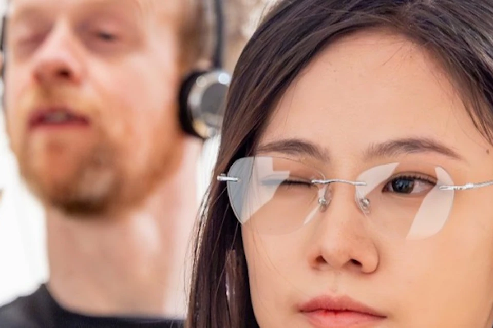 Two people wearing headphones, one with glasses, holding their hands up in a gesture, in a light indoor setting.