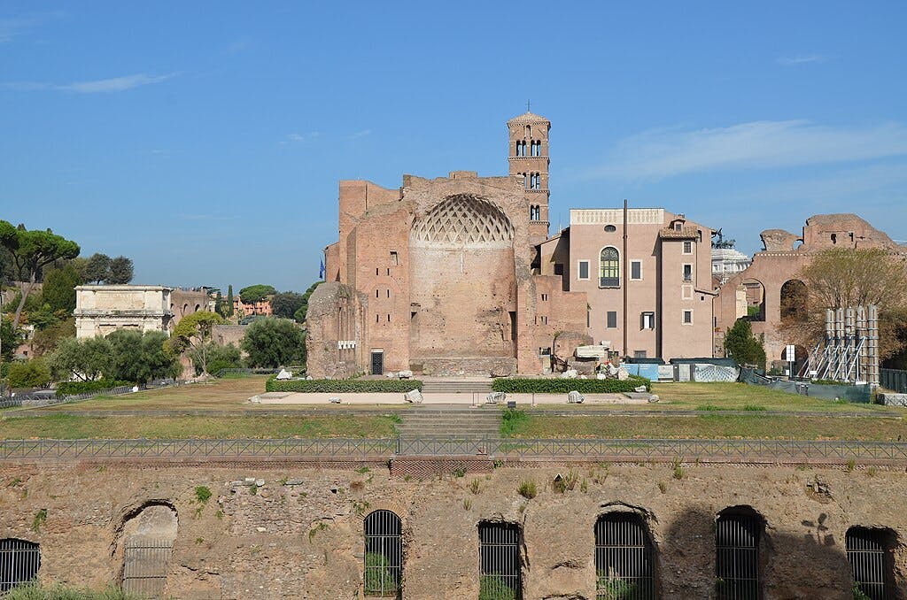 Uma vista das antigas ruínas romanas, com uma grande estrutura de tijolo com aberturas em arco e vegetação circundante sob um céu azul claro.
