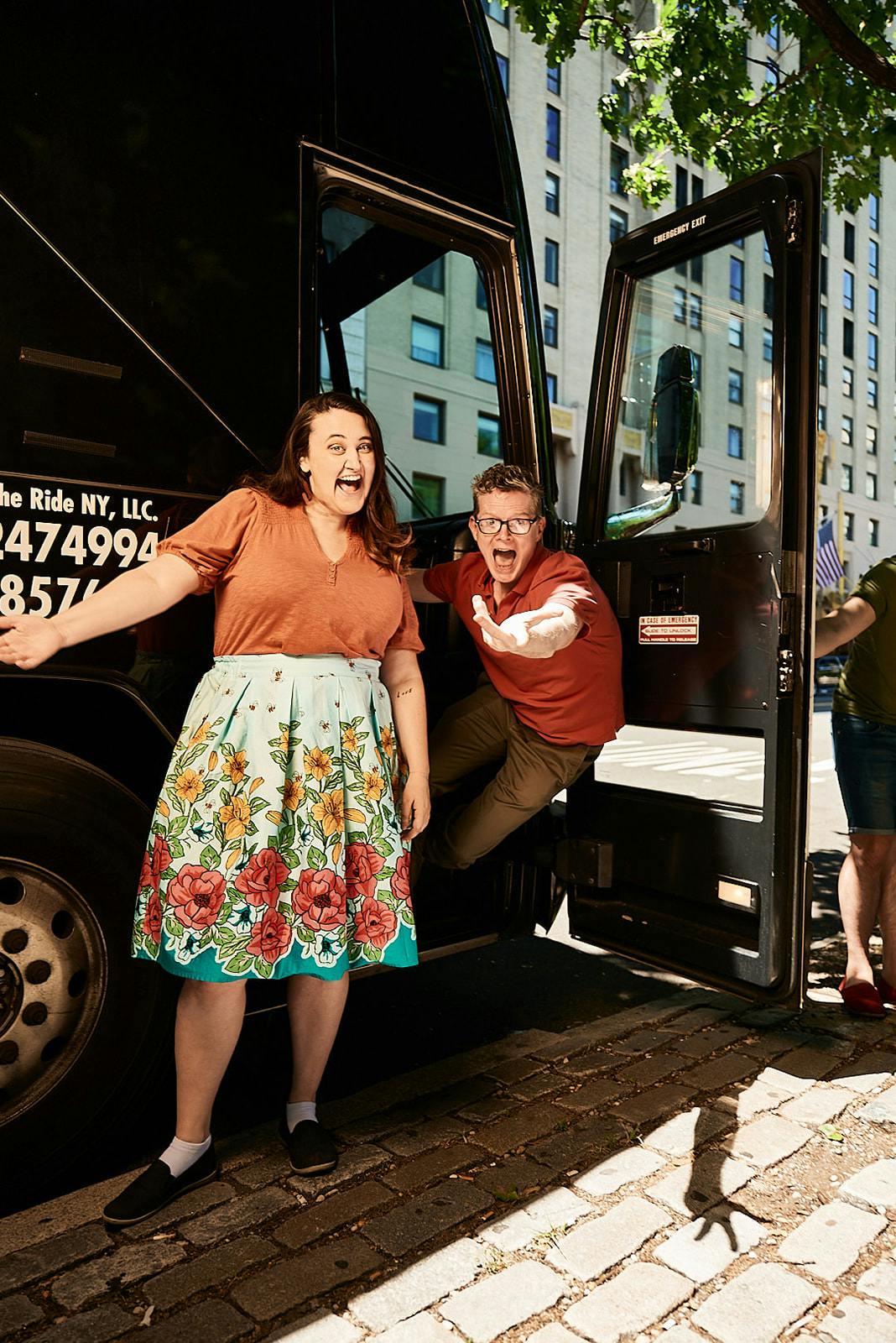 Δύο ενθουσιασμένοι άνθρωποι στέκονται δίπλα στην ανοιχτή πόρτα ενός μαύρου τουριστικού λεωφορείου σε μια αστική περιοχή με ψηλά κτίρια στο παρασκήνιο.