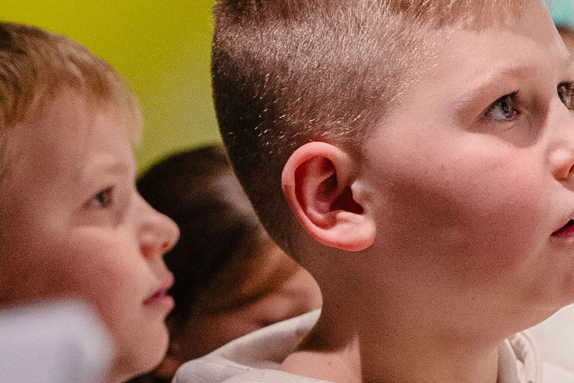 Children in hoodies are intently watching a display in a well-lit indoor exhibition, surrounded by people.