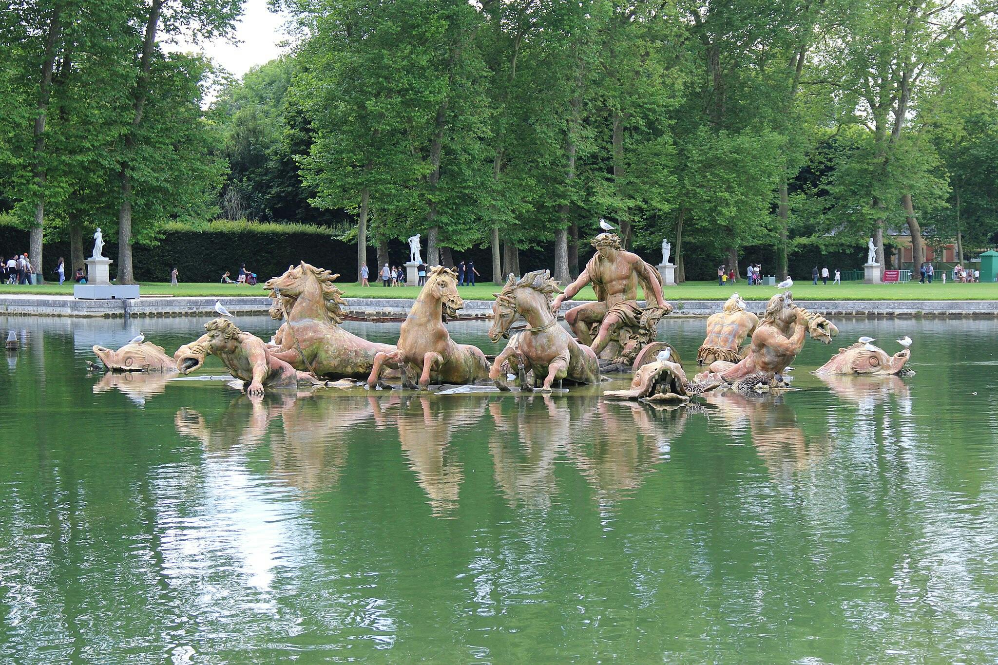 Фонтан со статуями лошадей и фигурами, выходящими из воды, в окружении деревьев и людей, идущих по тропинке на заднем плане.
