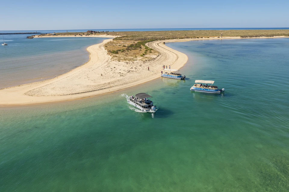 Fromosamar boats near a sandy island with clear green water, visitors walking on the beach under a sunny blue sky.